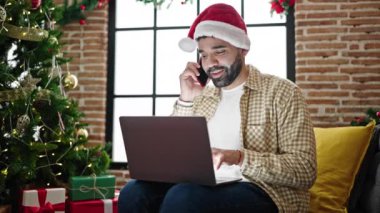 Genç İspanyol adam dizüstü bilgisayar kullanıyor. Akıllı telefonla konuşuyor. Noel 'i evde kutluyor.