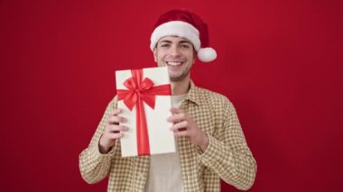 Genç İspanyol adam, kendine güveni tam, Noel şapkası takıyor ve kırmızı arka planda hediyesini tutuyor.