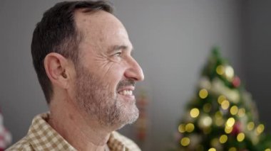 Orta yaşlı bir adam evde Noel ağacının yanında kendine güvenerek gülümsüyor.