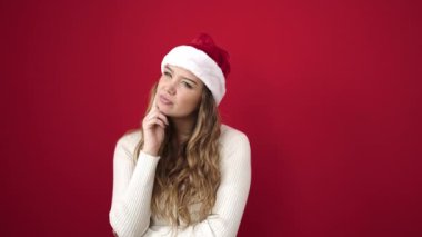 Noel şapkası takan genç ve güzel bir İspanyol kadın izole edilmiş kırmızı arka planda fikir ifadesiyle ayakta duruyor.