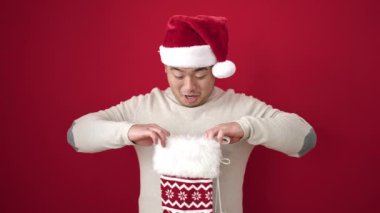 Genç Çinli adam gülümsüyor, kendine güveni tam, kırmızı arka planda Noel çorabı tutuyor.