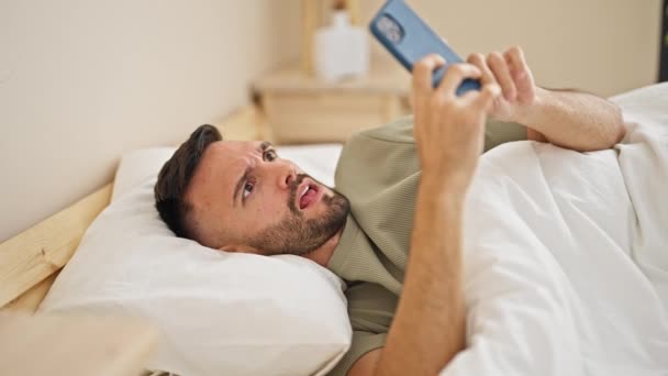 Ung Latinamerikan Man Med Smartphone Liggande Sängen Förvånad Över Sovrummet — Stockvideo