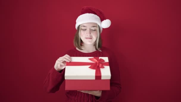 Młoda Blondynka Nosi Świąteczny Kapelusz Rozpakowując Prezent Odizolowanym Czerwonym Tle — Wideo stockowe
