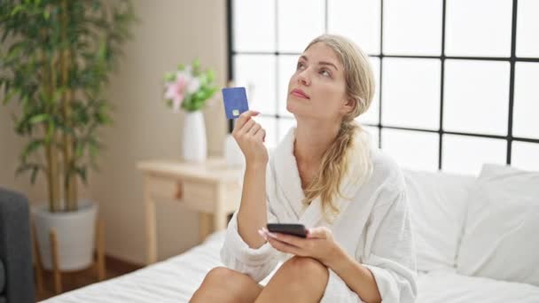 Young blonde woman wearing bathrobe shopping with credit card and smartphone thinking at bedroom