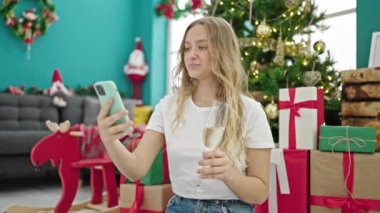Genç sarışın kadın Noel 'i kutluyor. Akıllı telefonuyla selfie çekiyor. Evde şampanya içiyor.