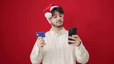Akıllı telefon ve kredi kartı kullanan genç İspanyol adam izole edilmiş kırmızı arka planda noel şapkası takıyor.