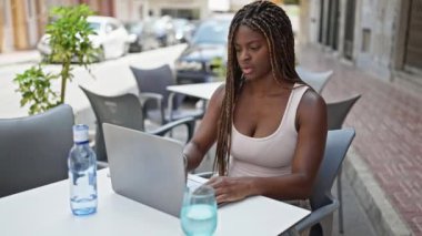 Kahve dükkanının terasında dizüstü bilgisayar kullanan Afrikalı Amerikalı kadın stresli bir şekilde oturuyor.