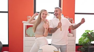 Kadın ve erkek, yemek odasında mutfak aletini mikrofon olarak kullanarak şarkı söyleyip dans ediyorlar.