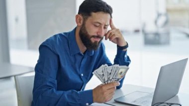 Genç İspanyol iş adamı ofiste dolar sayarak dizüstü bilgisayar kullanıyor.