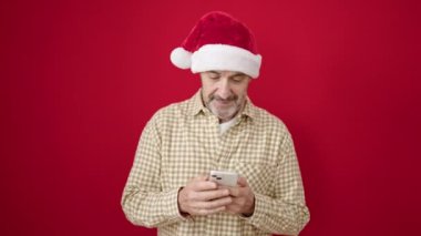 Yılbaşı şapkası takan orta yaşlı bir adam izole edilmiş kırmızı arka plan üzerinde akıllı telefon kullanıyor.