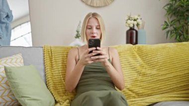 Akıllı telefon kullanan genç sarışın kadın evde stresli bir şekilde koltukta oturuyor.