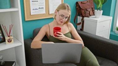 Genç sarışın iş kadını laptopunu ofiste kahve içerek kullanıyor.