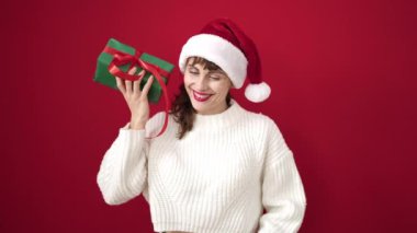 Kafkasyalı genç bir kadın Noel şapkası takarak gülümsüyor ve izole edilmiş kırmızı arka planda hediyesini dinliyor.