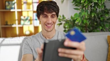 Genç İspanyol dokunmatik ped ve kredi kartıyla alışveriş yapıyor. Evdeki kanepede oturuyor.