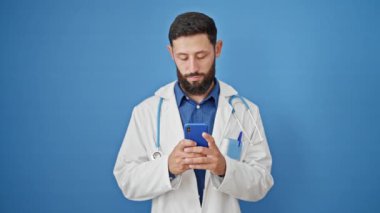 Genç İspanyol doktor mavi arka planda akıllı telefon kullanarak gülümsüyor.