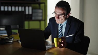 Orta yaşlı iş adamı ofiste kahve içerek dizüstü bilgisayar kullanıyor.