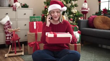 Young blonde woman talking on smartphone sitting on floor by christmas tree at home