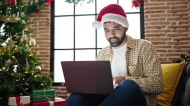 Evde dizüstü bilgisayar kullanan genç İspanyol adam Noel şapkası takıyor.