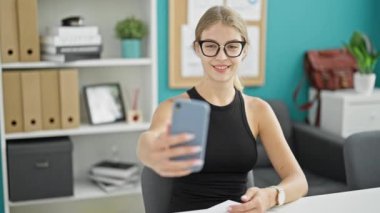Genç sarışın iş kadını ofiste gülümseyen akıllı telefonla selfie çekiyor.