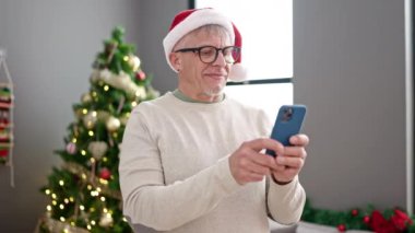 Orta yaşlı, kır saçlı, akıllı telefon kullanan bir adam Noel ağacının yanında duruyor.