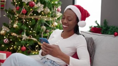 Afrika kökenli Amerikalı kadın akıllı telefon kullanıyor. Yılbaşı ağacının yanında oturuyor.