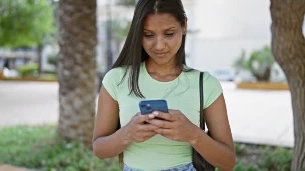 公園でスマートフォンを笑顔に使っている若いラテン系女性 — ストック動画