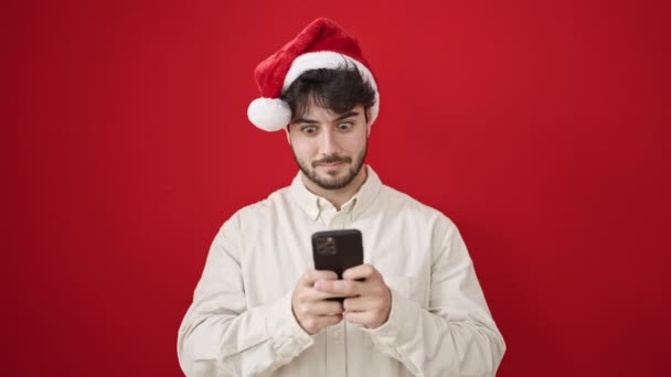 Junger Hispanischer Mann Trägt Weihnachtsmütze Mit Smartphone Vor Isoliertem Roten — Stockvideo