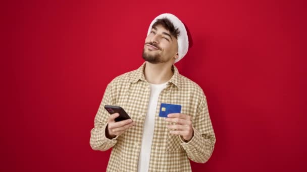 年轻的Arab男子用智能手机在孤立的红色背景下进行圣诞网上购物 — 图库视频影像