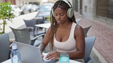 Laptop ve kulaklık kullanan Afrikalı Amerikalı bir kadın kahve dükkanının terasında bir bardak su içiyor.