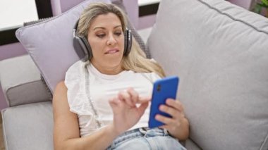 Genç sarışın kadın akıllı telefon kullanıyor. Kulaklık takıyor. Evdeki kanepede yatıyor.