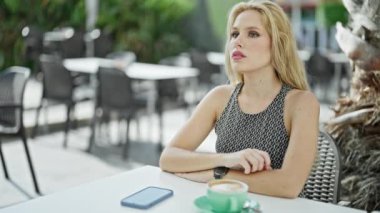 Akıllı telefon kullanan genç sarışın kadın kahve dükkanının terasına üzgün üzgün bakıyor.