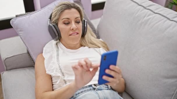 年轻的金发女人带着耳机用智能手机躺在家里的沙发上 — 图库视频影像