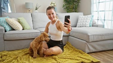 Köpekli beyaz bir kadın evde yerde oturmuş akıllı telefonuyla selfie çekiyor.