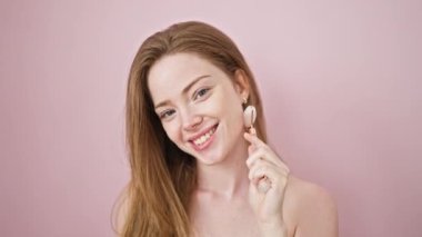 Genç, sarışın bir kadın. Yüzüne masaj yapıyor. Teninde pembe bir arka plan var.