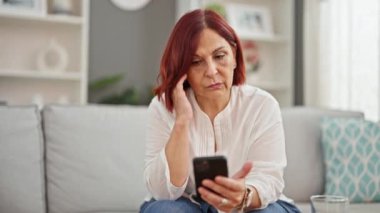 Orta yaşlı bir kadın evde ciddi bir ifadeyle akıllı telefon kullanıyor.