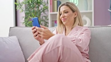 Evdeki kanepede oturan akıllı telefon kullanan genç sarışın kadın.