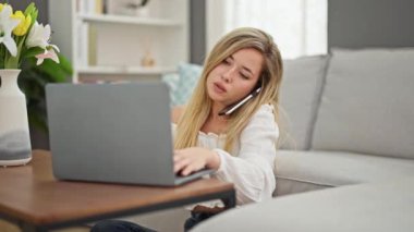 Evde dizüstü bilgisayar kullanan genç sarışın kadın akıllı telefondan konuşuyor.
