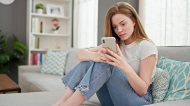 Akıllı telefon kullanan genç sarışın kadın evde üzgün görünüyor.