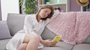 Kanepede oturan akıllı telefon kullanan genç sarışın kadın ciddi bir yüzle evde oturuyor.