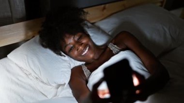 Afrika kökenli Amerikalı kadın iç çamaşırı giyiyor. Yatak odasında yatarken akıllı telefon kullanıyor.
