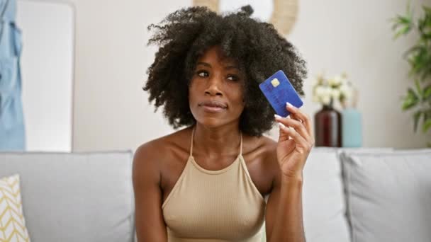 拿着信用卡坐在沙发上想着家里的非裔美国女人 — 图库视频影像