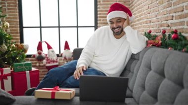 Noel Baba şapkalı mutlu adam Noel 'de oturma odasında dizüstü bilgisayarında video görüşmesi yapıyor.