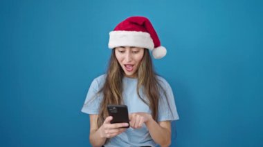 Noel Baba şapkalı genç bir kadın mavi arka planda akıllı telefonuyla selfie çekiyor. Noel mevsimi kutlaması