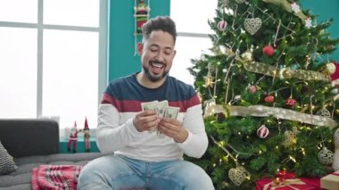 Yakışıklı, sakallı, süveterli genç adam Noel zamanı oturma odasında para sayıyor.