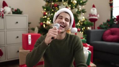 Oturma odasında Noel ağacıyla şarkı söyleyen mutlu yakışıklı genç adam.