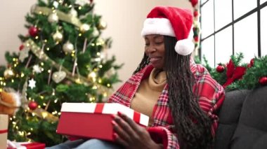 Afrikalı bir kadın hediye paketini açıyor Noel ağacının yanında oturuyor.