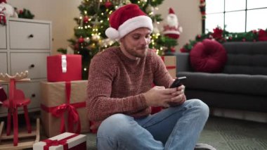 Noel Baba şapkalı mutlu genç adam Noel 'de evde akıllı telefon kullanıyor.