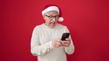Noel Baba şapkalı ve kazaklı heyecanlı adam akıllı telefon kullanıyor, kırmızı arka plandaki başarıyı kutluyor.