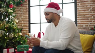 Noel Baba şapkalı genç adam Noel 'de koltukta cep telefonu kullanıyor.