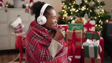 Gülümseyen genç bir kadın, Noel arka planında kulaklıkla müzik dinlerken kahve içiyor.